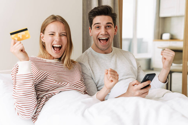 Portrait of beautiful excited couple holding smartphone and credit card while making winner gesture in bed - Φωτογραφία, εικόνα