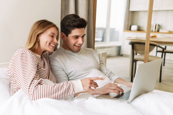 Portrait of beautiful joyful couple smiling and using laptop while lying in bed at home - Fotó, kép