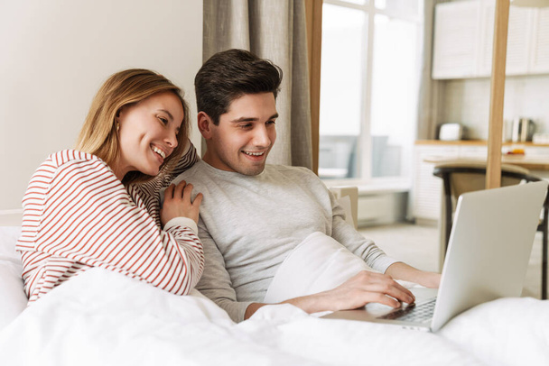 Portrait of beautiful joyful couple smiling and using laptop while lying in bed at home - Φωτογραφία, εικόνα
