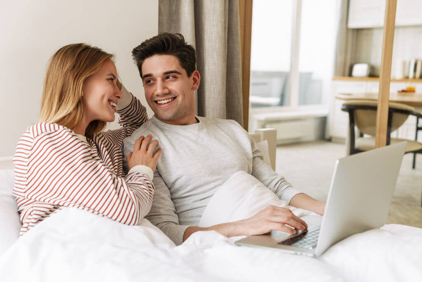 Portrait of beautiful joyful couple smiling and using laptop while lying in bed at home - 写真・画像