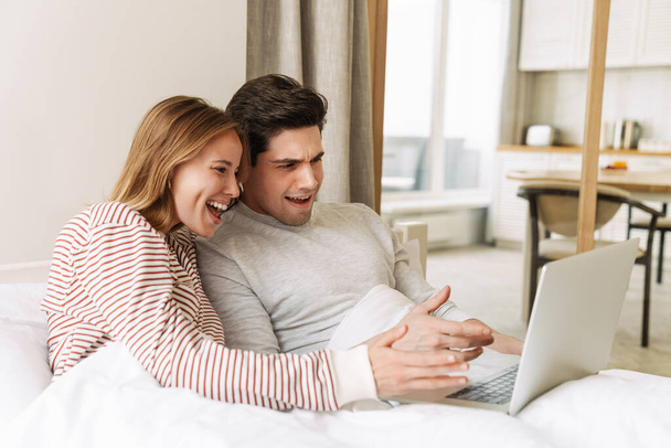 Portrait of beautiful excited couple smiling and using laptop while lying in bed at home - Photo, Image