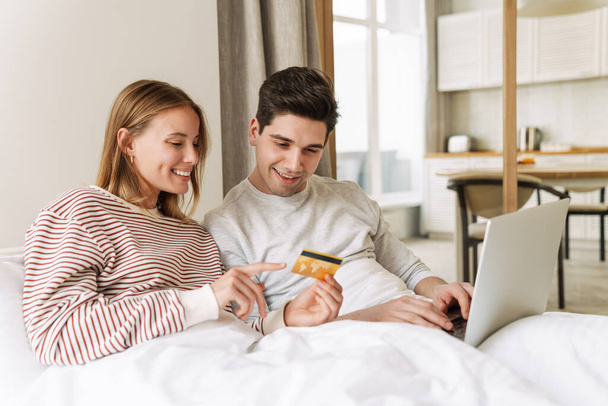 Portrait of beautiful joyful couple holding credit card and using laptop while lying in bed at home - Fotoğraf, Görsel