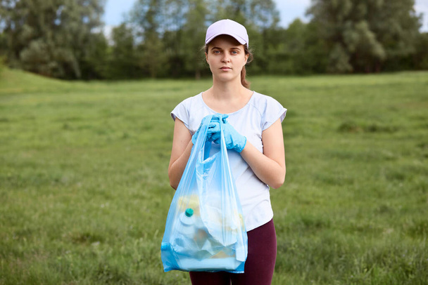 Young woman in white t shirt and baseball cap holding blue garbage bag in hands, female collecting litter in meadow, looks at camera, stands against trees on background. - Фото, зображення