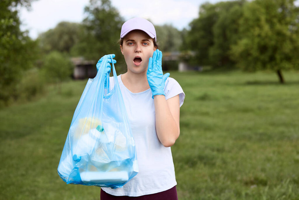 Astonished girl with widely opened mouth stands with garbage bag in hand, keeping palm on her cheek, being shocked of planet pollution, dresses casual clothing and baseball cap. - Photo, Image