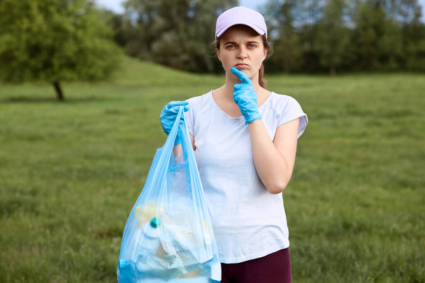 Pensive woman in baseball cap and casual clothing posing with garbage bag, looking at camera and keeps hand on chin, finds solution of pollution problems. - Φωτογραφία, εικόνα