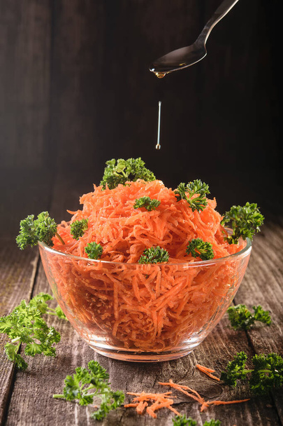 Измельченная морковь с петрушкой в прозрачной миске на темном деревянном фоне, ложка с оливковым маслом, вегетарианская концепция здорового питания. - Фото, изображение