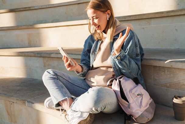 Beautiful smiling young blonde woman wearing denim jacket sitting on steps outdoors, talking via mobile phone and earphones - Photo, image