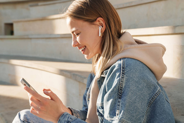 Beautiful smiling young blonde woman wearing denim jacket sitting on steps outdoors, listening to music with earphones and mobile phone - Foto, immagini