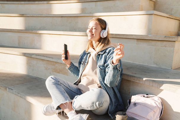 Beautiful smiling young blonde woman wearing denim jacket sitting on steps outdoors, listening to music with headphones and mobile phone - Photo, Image