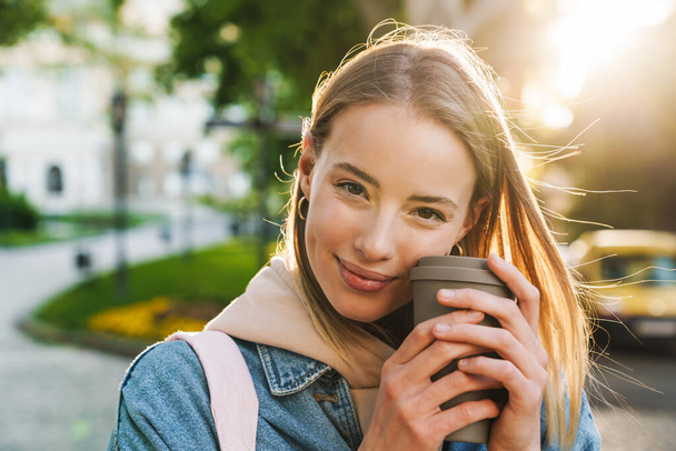 Beautiful cheerful young blonde woman wearing denim jacket walking in the city, holding takeaway coffee cup - Foto, imagen