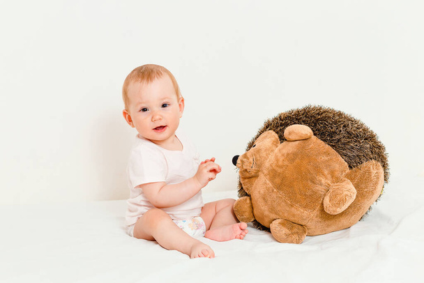 Pequeno bebê brinca com um grande brinquedo de ouriço macio sentado na cama no fundo branco
 - Foto, Imagem