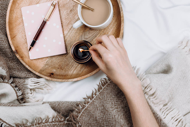 Cozy flatlay with wooden tray, cup of coffee or cocoa, candle, notebooks on white sheets and blankets - Fotó, kép