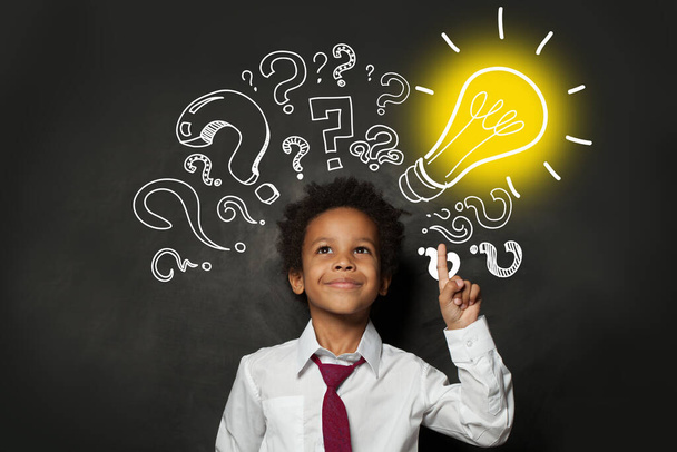 Smiling black child student with lightbulb on blackboard background. Brainstorming and idea concept - Photo, Image