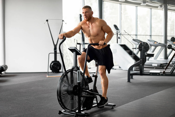 Man exercise bike gym cycling training fitness. Fitness male using air bike cardio workout. Athlete guy naked torso biking indoor gym exercising his legs. Cross functional training. - Photo, image