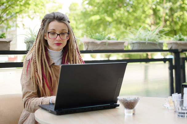Urocza kobieta przedsiębiorca w okularach z długimi dredami casual ubrany planuje proces pracy przy użyciu aplikacji na laptopie podczas siedzenia w nowoczesnej kawiarni ulicznej na świeżym powietrzu - Zdjęcie, obraz
