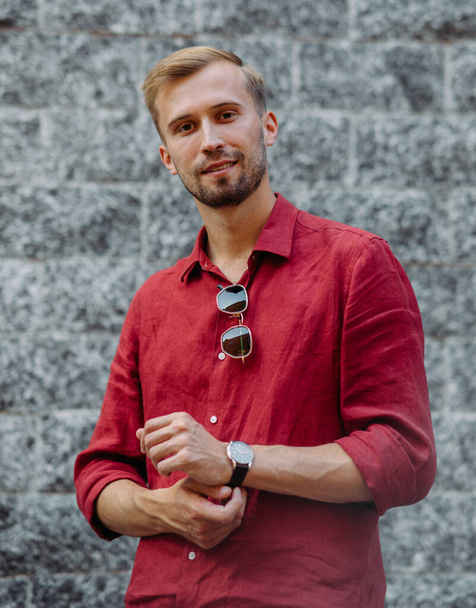 Portrait of young man in red shirt with sunglasses against background of grey brick wall. - Photo, Image