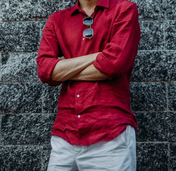 Young man in red shirt and white pants with sunglasses stands against background of grey brick wall. Closeup. - Photo, Image