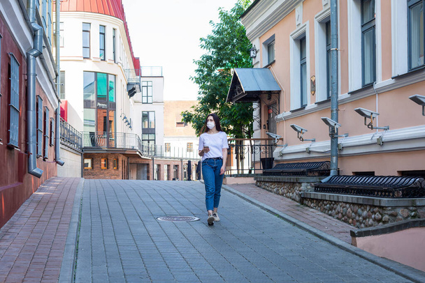 Caucasian young woman in a protective mask on her face walks alone along the street, listens to music. Concept: solo walks great outdoors in uninhabited places. Europe. City environment. Summer morning - Foto, Imagen