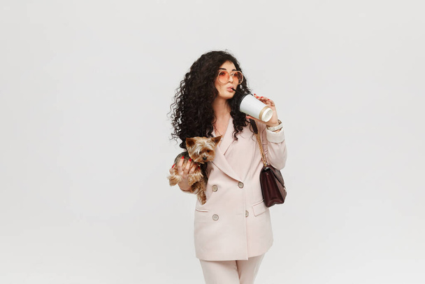 A young woman in a beige suit holding Yorkie terrier in the hand and drinking a coffee, isolated with copy space. - Fotografie, Obrázek