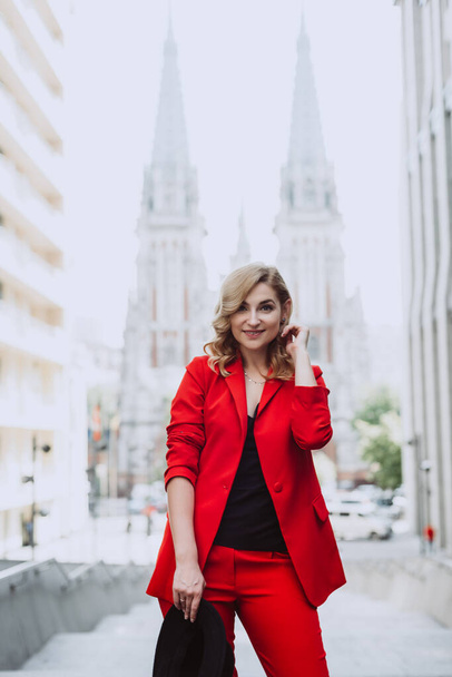 Young beautiful blond woman in a red suit and black hat stands outdoors in the yard next to the building. Stylish people. Soft selective focus. - Photo, Image