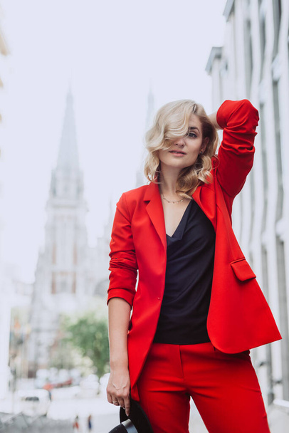Young beautiful blond woman in a red suit and black hat stands outdoors in the yard next to the building. Stylish people. Soft selective focus. - 写真・画像
