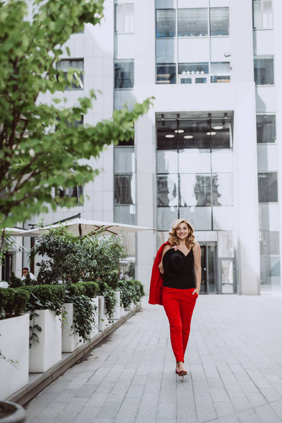 Νεαρή επιχειρηματίας ξανθιά με κόκκινο κοστούμι είναι το περπάτημα σε εξωτερικούς χώρους στην αυλή ενός επιχειρηματικού κέντρου. Κομψοί άνθρωποι. Μαλακή επιλεκτική εστίαση. - Φωτογραφία, εικόνα