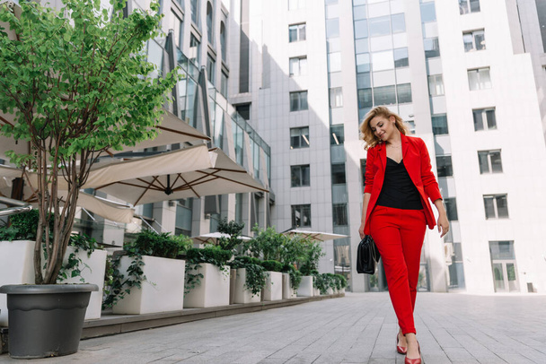 ビジネスセンターの中庭では、赤いスーツの若いビジネス女性ブロンドが屋外を歩いています。スタイリッシュな人々。柔らかい選択的フォーカス. - 写真・画像