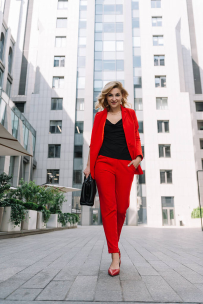 ビジネスセンターの中庭では、赤いスーツの若いビジネス女性ブロンドが屋外を歩いています。スタイリッシュな人々。柔らかい選択的フォーカス. - 写真・画像