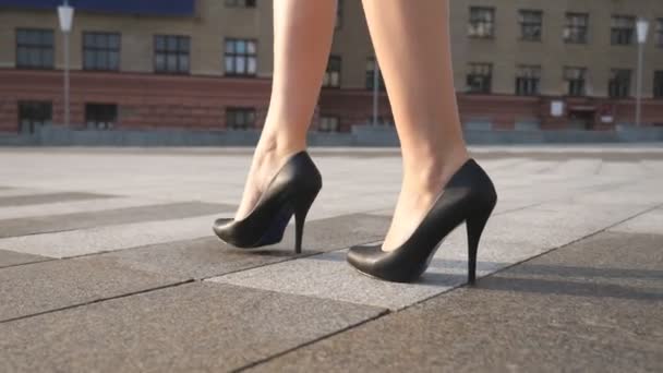 Slim gambe femminili in scarpe nere sui tacchi alti a piedi in piazza della città in una giornata di sole. Piedi di donna d'affari giovane in calzature su tallone alto che va in strada urbana. Vista ad angolo basso Movimento lento Avvicinamento - Filmati, video