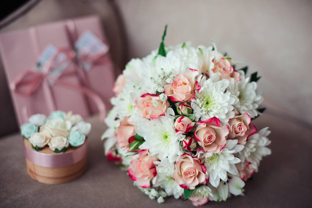 Крупный план свадебного букета из розовых роз и белых хризантем на фоне розовых сертификатов. Коробка для колец на пушистом диване
 - Фото, изображение