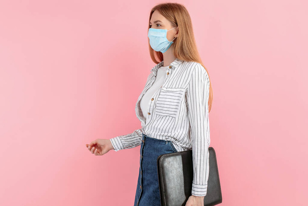 Image of a happy young business woman in a medical protective mask on her face, standing with a folder of documents on an isolated pink background - Photo, image