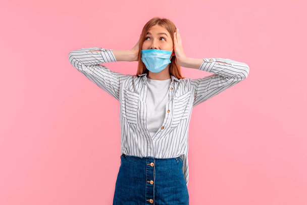 angry irritated girl in a medical protective mask on her face, angrily frowns and covers her ears with her hands isolated on a pink background - 写真・画像