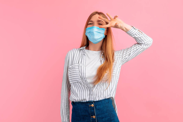 image of a cheerful young woman wearing a medical protective mask on her face, dressed in casual clothing, smiling and showing a peace sign with two fingers isolated on a pink background in the Studio - Foto, imagen