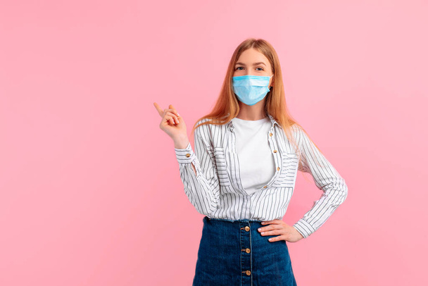 attractive young woman in a medical protective mask on her face, points her finger at copyspace, on an isolated pink background - Foto, immagini