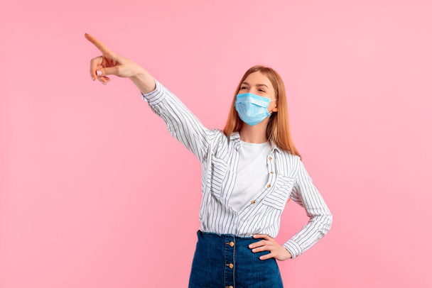young woman in a medical protective mask on her face, pointing into the distance, notices something amazing on a pink background - Фото, изображение