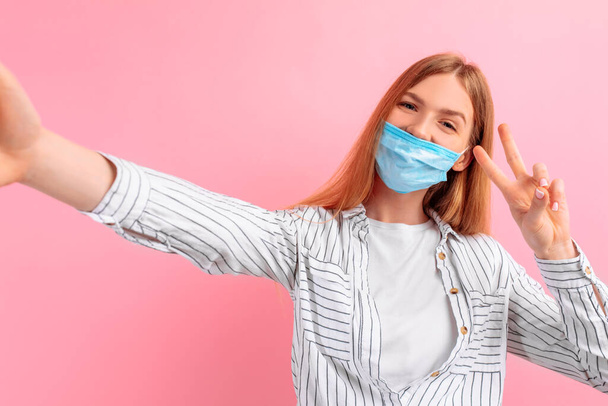 girl in a medical protective mask takes a selfie on her smartphone and shows a peace gesture with two fingers, on a pink background. The epidemic of coronavirus - Foto, immagini