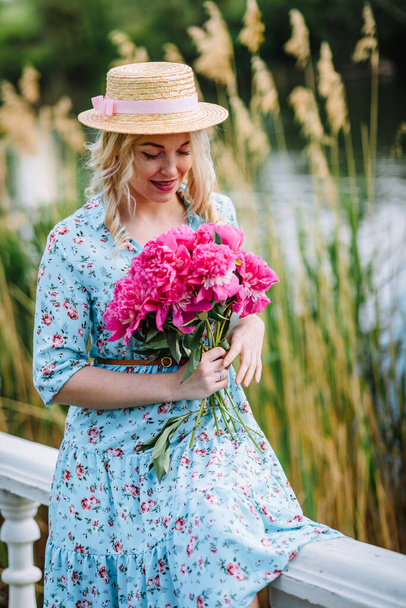 ピンク色の牡丹の花束を持つ若い女性が池のビーチに休んでいます。青いドレスの女の子と花の帽子。屋外レクリエーションの概念。夏休み. - 写真・画像