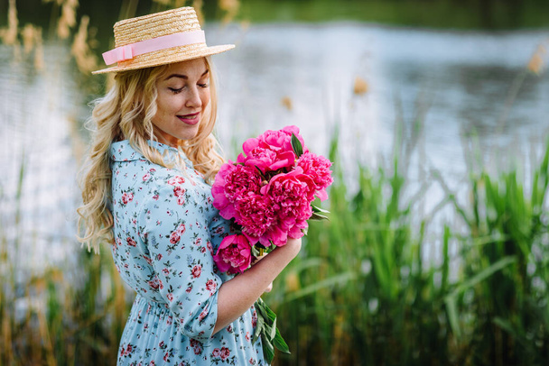 Een jonge vrouw met een boeket roze pioenrozen rust op een vijverstrand. Meisje in een blauwe jurk en een hoed met bloemen. Het concept van recreatie buitenshuis. Zomervakantie. - Foto, afbeelding