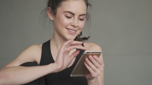 A close-up view of a beautiful young woman in a black tracksuit is using her smartphone before training in a gray studio in the morning - Video, Çekim