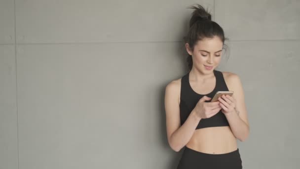 A charming young woman in a black tracksuit is using her smartphone standing near the wall before training in a gray studio in the morning - Video, Çekim