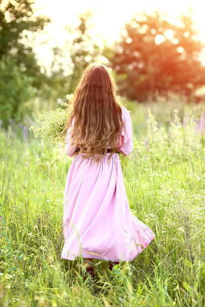 Young beautiful red-haired girl in a pink dress in the garden at sunset. The effect of bokeh, flash, sun rays. Summer. People and nature concept. - Photo, Image