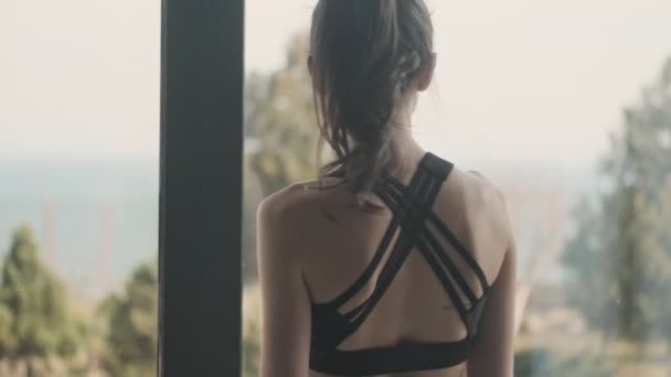 A back view of a slim calm young woman in a black tracksuit is standing near window in a gray studio - Metraje, vídeo