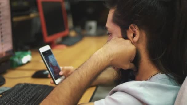 Homme hindou avec dreadlocks et une barbe est assis à un ordinateur dans le bureau et regarde le smartphone. - Séquence, vidéo