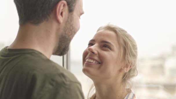 A happy young couple in love are hugging and kissing while standing near window at home - Filmagem, Vídeo