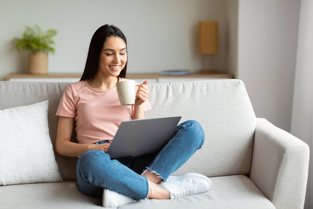 Mulher feliz usando laptop desfrutando de café sentado no sofá interior
 - Foto, Imagem