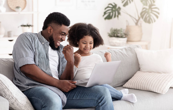 Heyecanlı siyah baba ve küçük kızı evde dizüstü bilgisayarla zaferi kutluyor. - Fotoğraf, Görsel