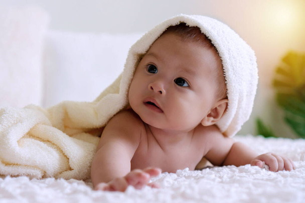 Χαρούμενο χαριτωμένο μωρό ξαπλωμένο κάτω από λευκή κουβέρτα κοιτάζοντας κάτι. Αθώο μωρό σέρνεται στο λευκό κρεβάτι με πετσέτα στο κεφάλι του στο σπίτι. - Φωτογραφία, εικόνα