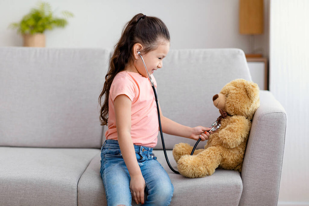 Menina jogando médico com ursinho de pelúcia sentado no sofá dentro de casa
 - Foto, Imagem