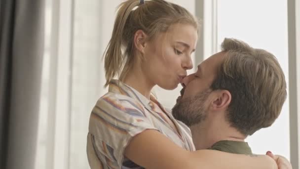 A close-up view of a pretty nice young couple man and woman are hugging and kissing while standing near window at home - Materiał filmowy, wideo