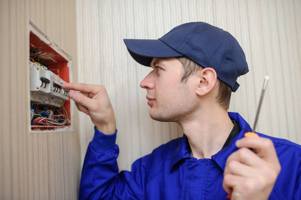 πλευρική άποψη ενός νεαρού Eletrician σε μπλε συνολικά αποσυναρμολόγηση ηλεκτρικού πίνακα με ασφάλειες σε hous - Φωτογραφία, εικόνα